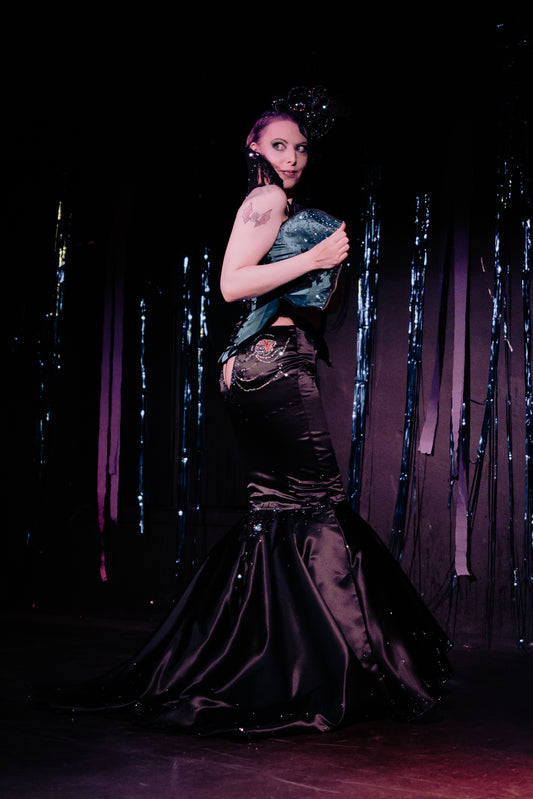 Burleskiesiintyjä seisoo lavalla avaten korsettiaan, hymyilee olan yli yleisölle.